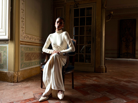 Woman in cream-colored clothing seated in an ornate period room with wooden doors and decorative wall panels.