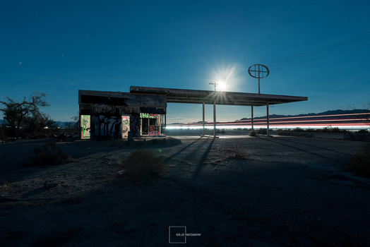 Abandoned gas station at dusk with glowing interior lights and motion-blurred vehicle light trails passing by.