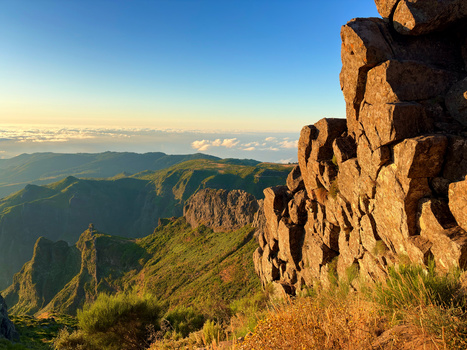 Rocky cliff edge overlooking a vast valley with forested slopes and distant mountains under clear blue sky.