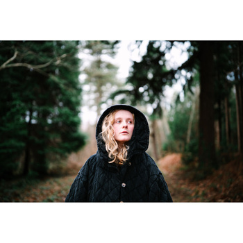 Young child in black coat and hood looking upward in a tree-lined forest path.
