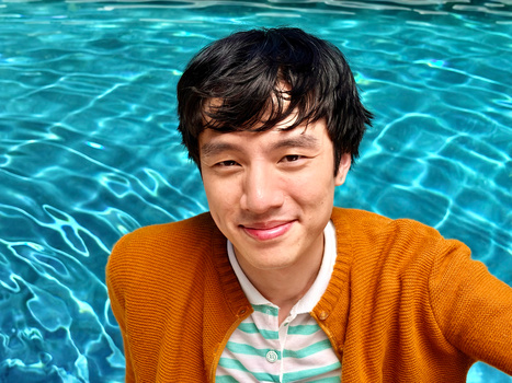 Young person in orange cardigan smiling at camera poolside with turquoise water behind.