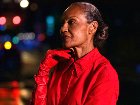 Woman in red jacket looking over shoulder against blurred nighttime city lights.