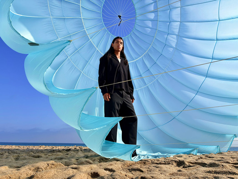 Woman in dark clothing stands within a massive light blue parachute canopy against a desert landscape.