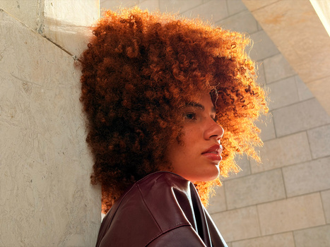Person with voluminous curly copper-colored hair in profile against white tiled wall, wearing burgundy jacket.