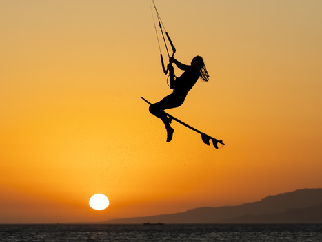 Silhouette of a kitesurfer airborne against an orange sunset over water.