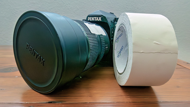 Pentax camera lens with protective lens cap and roll of white tape on wooden surface.