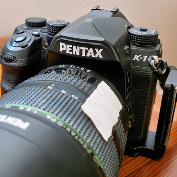 Pentax K-7 DSLR camera with attached lens on wooden surface, shot from above angle.