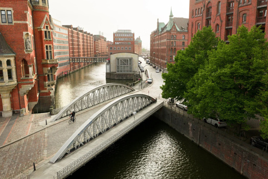 Arched pedestrian bridge spanning a canal with red brick buildings lining the waterway in an urban European city.