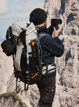 Photographer wearing a large backpack and beanie adjusts a telephoto lens while standing against a rocky cliff backdrop.