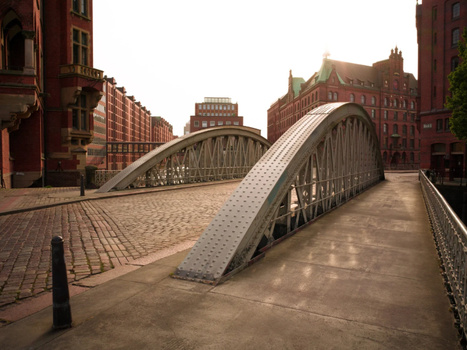 Arched pedestrian bridge with metal railings spanning a brick plaza between red brick buildings.