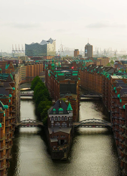 Aerial view of a historic European city with red-brick buildings, waterways, and modern towers in the distance.