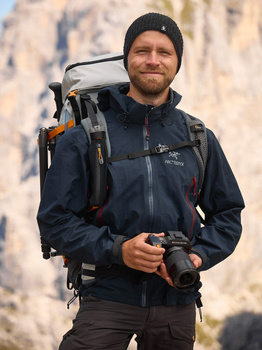 Male photographer wearing dark jacket and beanie holds mirrorless camera while carrying backpack with climbing gear against rocky cliff backdrop.