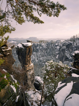Snow-dusted rock outcropping frames a winter landscape of forested valleys and distant mountains.