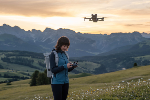 Drone operator holding remote controller while flying quadcopter over alpine meadow at sunset.