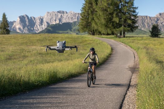 Cyclist riding on a paved path through a grassy valley with mountains and evergreen trees in the background.