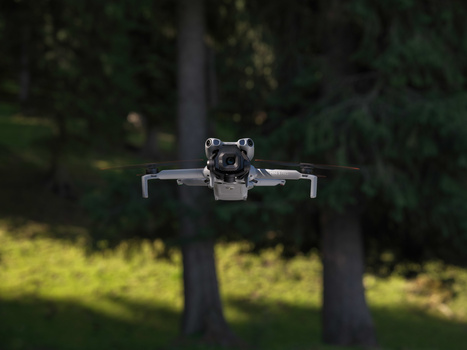 Compact quadcopter drone hovering in midair with forest trees blurred in background.