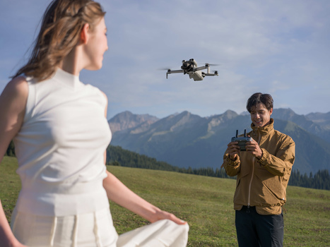 Operator flying a quadcopter drone while two people stand in an alpine meadow with mountains in the background.