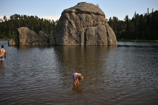 Large granite boulder in a lake surrounded by coniferous forest, with swimmers in the foreground.