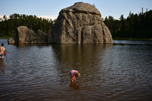 People wading in a lake with massive granite boulders and coniferous forest in the background.