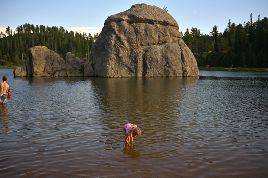 Large rounded boulder rising from a calm lake surrounded by evergreen forest, with swimmers in the shallow water.