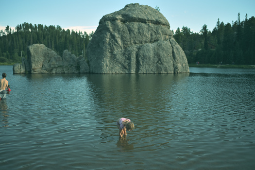Two swimmers in a lake with a large granite boulder formation and evergreen forest in the background.