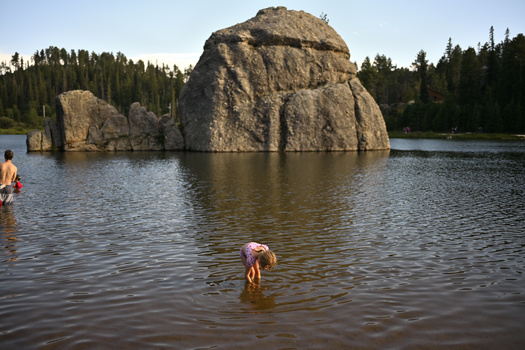 Large rounded boulder rising from lake water with swimmers in foreground and forested shoreline behind.