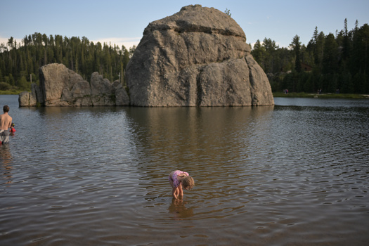 People wading in a lake with a massive rounded granite boulder and forested shoreline in the background.