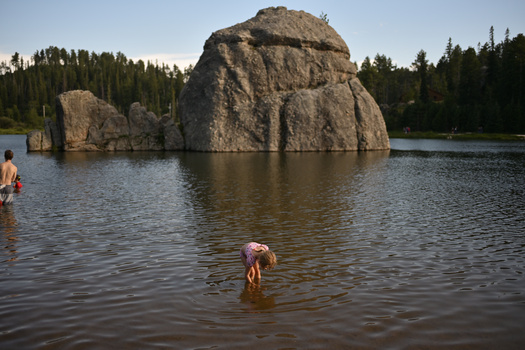 Swimmers in a lake with a massive rounded boulder formation rising from the water, surrounded by coniferous forest.