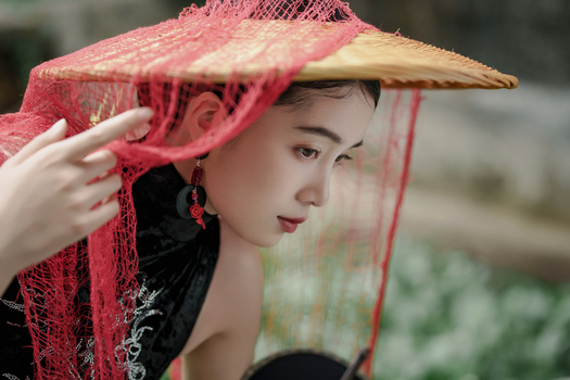 Woman wearing a pink conical straw hat with red mesh veil, adjusting the fabric with her hand.