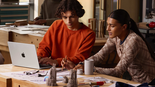 Two people collaborating at a wooden desk while reviewing work on a laptop and architectural models.