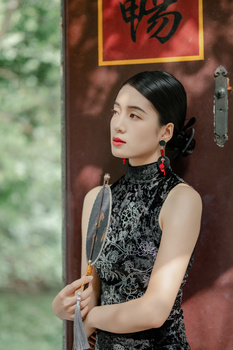 Woman in black patterned sleeveless dress with red lipstick posed against warm-toned wooden background with Asian characters.