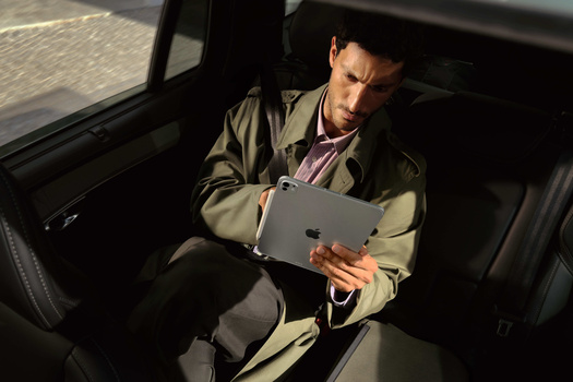 Man in tan jacket sits in car's back seat reviewing content on tablet device.
