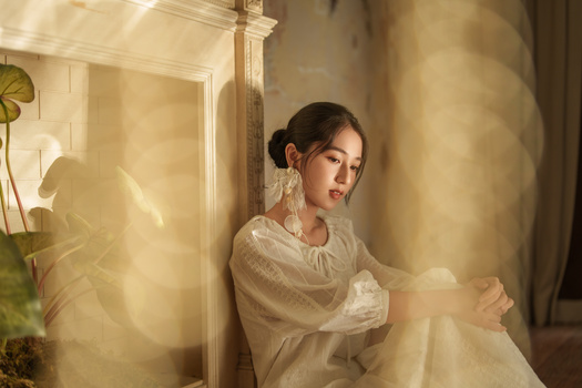 Woman in cream linen shirt posed against warm-toned wall with soft window light and sheer fabric overlay.