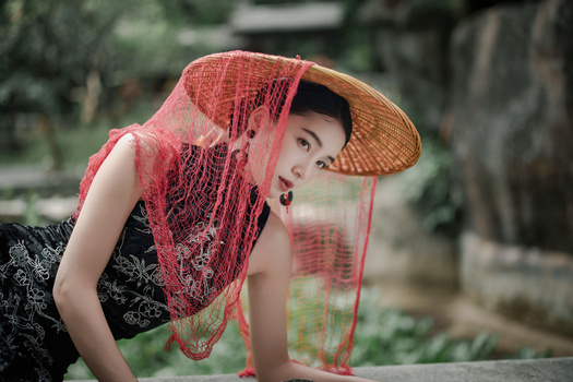 Woman in traditional conical hat draped with red mesh fabric, posed against blurred garden background.