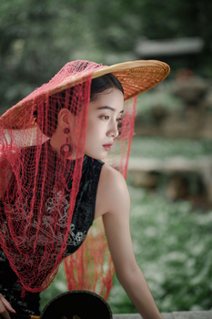 Woman wearing a traditional conical hat with red mesh netting draped over her face and shoulders.