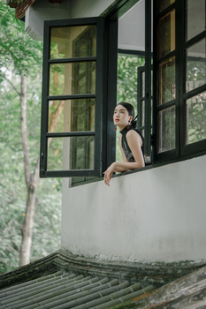 Woman leaning out of a black-framed window in a white-walled building surrounded by green foliage.