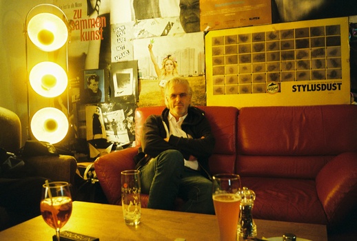 Man in dark blazer seated on red couch in studio lit by three circular yellow lights.