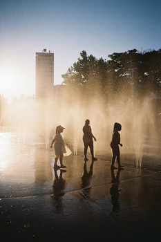 Three surfers in wetsuits standing in shallow water surrounded by sea spray and mist, with a tall building visible in the foggy background.