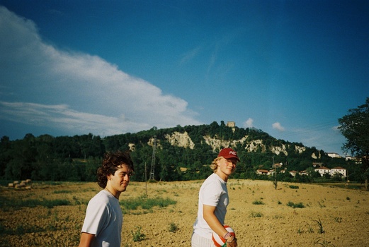 Two young men walking across an open field with a hillside town visible in the distance.