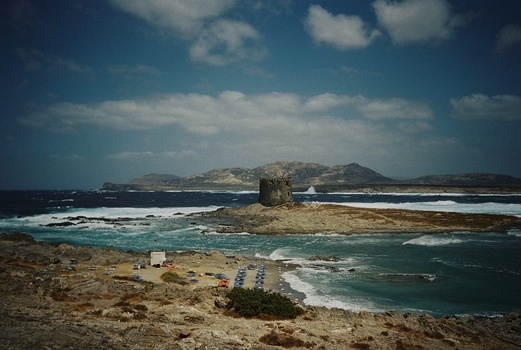Rocky coastal landscape with a solitary stone tower on a small island surrounded by turquoise waters.