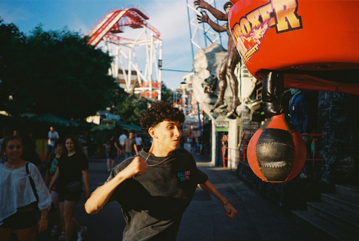Young man in black shirt jogging past amusement park rides during golden hour.