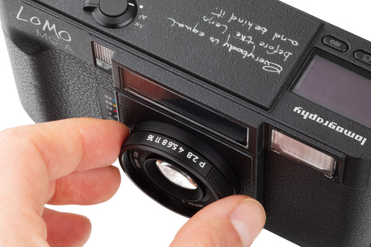 Hand holding the lens of a compact rangefinder camera, demonstrating the camera's construction and focusing mechanism.