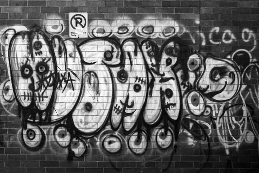 Black and white photograph of elaborate graffiti tags on a brick wall with overlapping letters and circular elements.