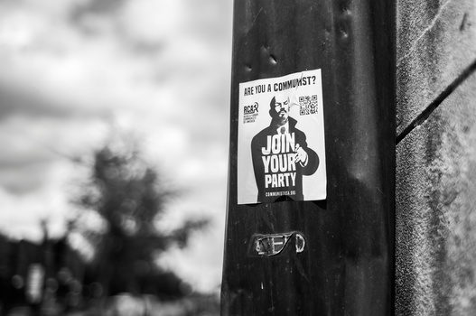 Black and white photograph of a political recruitment sticker on a dark pole with blurred background.