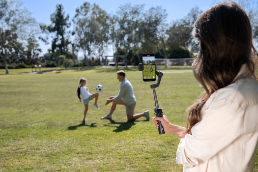 Woman filming children playing soccer on a grass field using a smartphone mounted on a gimbal stabilizer.
