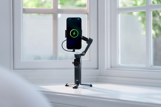 Smartphone mounted on a three-axis gimbal stabilizer displaying a charging indicator on its screen, positioned on a bright windowsill.