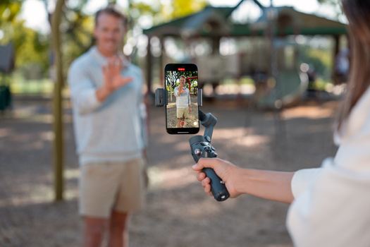 Person holding a smartphone on a gimbal stabilizer, recording video of a man in casual clothing standing in a park.
