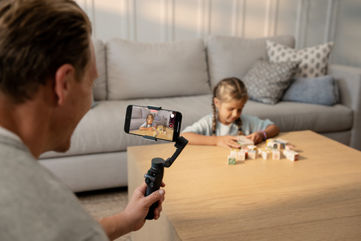 Man holding smartphone on gimbal stabilizer while recording child playing with building blocks at wooden table.