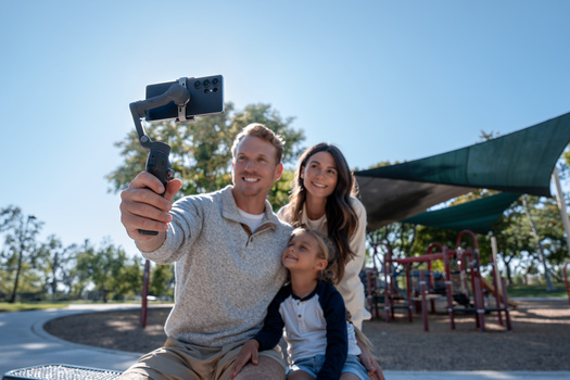 Family of three taking a selfie at a playground on a sunny day.