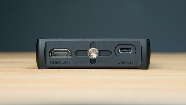 Close-up of a black device connector panel showing HDMI OUT, USB 3.0, and a central button on a wooden surface.
